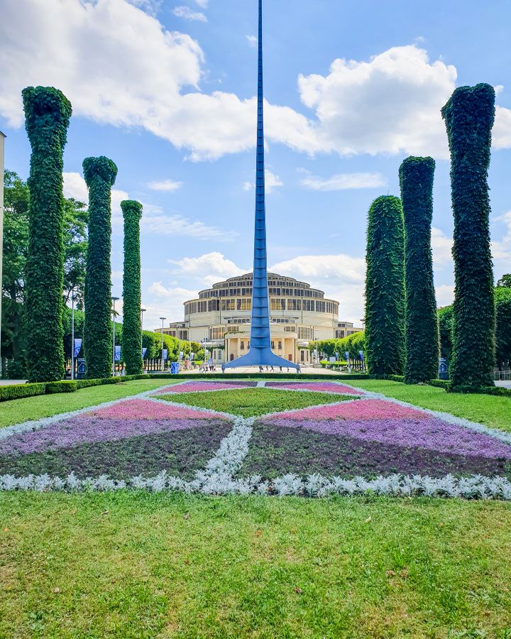Poland Szczytnicki Park How to Have a Wonderful Weekend in Wrocław on a Budget A giant metal spire in front of a big round building in Szczytnicki park in Wrocław, Poland. In front of the spire is a flower bed with purple and pink coloured flowers. On either side are big structures covered in green plants. "How to Have a Wonderful Weekend in Wrocław on a Budget"