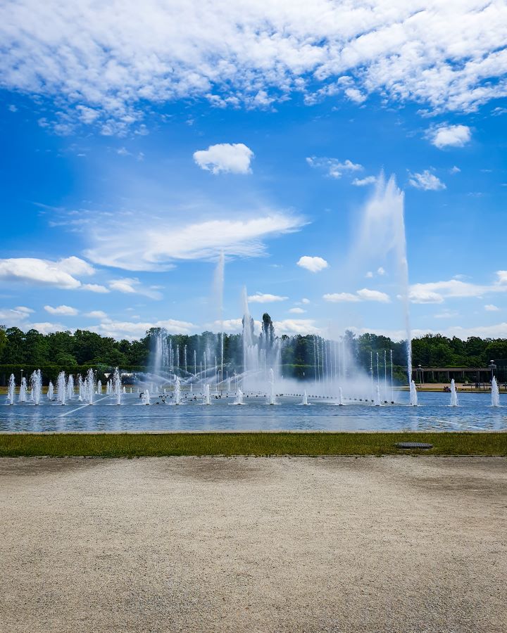 Poland Szczytnicki Park How to Have a Wonderful Weekend in Wrocław on a Budget Some brown dirt and grass with a blue body of water behind it. There's lots of fountain jets coming out of it and behind that are trees and the blue sky in Wrocław, Poland. "How to Have a Wonderful Weekend in Wrocław on a Budget"