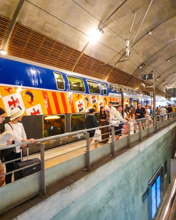 A double decker train at a station in Monaco. There's people on the train and on the platform and the platform is indoors under a curved concrete roof. "The Best Budget Nice to Monaco Day Trip"