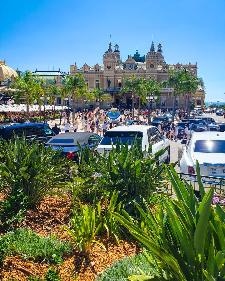 In the foreground are green plants and fancy cars parked outside the Monte-Carlo casino in Monaco. The casino is a big sand coloured building with clock towers and there's also palm trees out the front. There are also lots of people wandering around and above is a clear blue sky. "The Best Budget Nice to Monaco Day Trip"