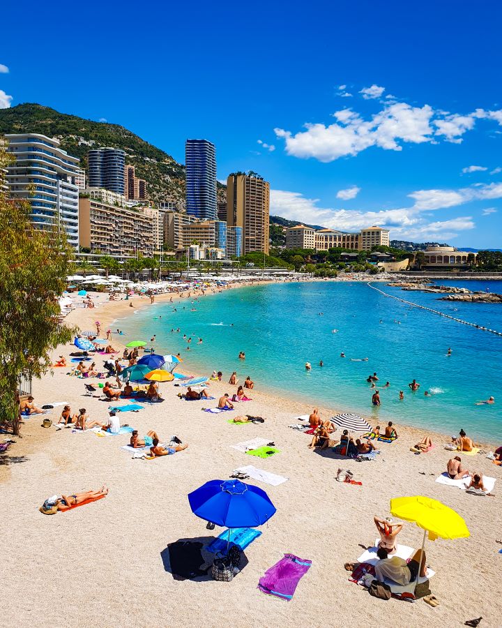 The beautiful Larvotto beach in Monaco. There's the sand on the left with lots of tall buildings behind it. On the right is the clear blue water and there's lots of people everywhere enjoying the sun. "The Best Budget Nice to Monaco Day Trip"