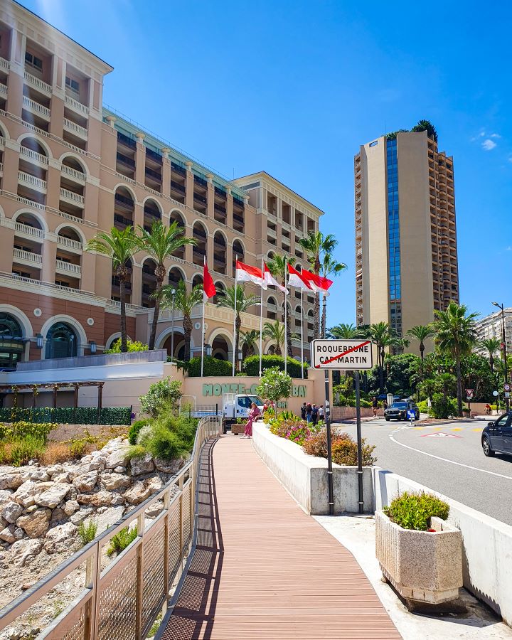 A border from France to Monaco with five Monaco flags on poles which is red on the top and white on the bottom. In front of the flags is a path, some rocks on the left and a road on the right. Behind the flags are some big hotels and the blue sky. "The Best Budget Nice to Monaco Day Trip"