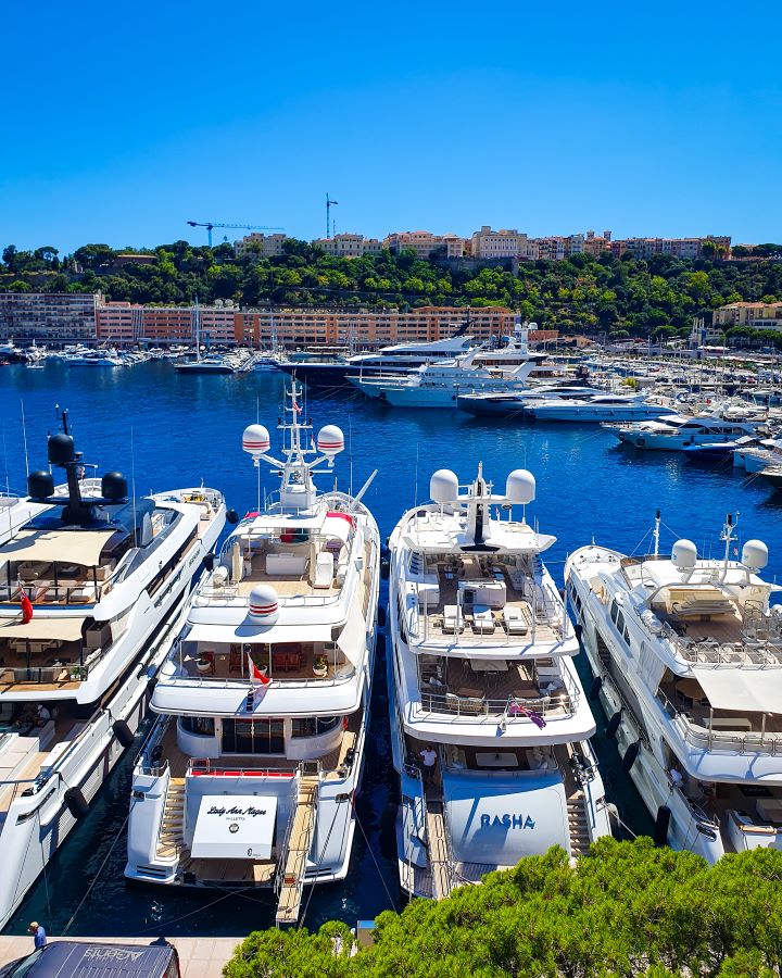 The Best Budget Nice to Monaco Day Trip. Looking out over Monaco harbour to lots of big white yachts. Behind them is a hill with green trees and orange and yellow buildings on top in Monaco City. The sky and harbour water are a beautiful clear blue.