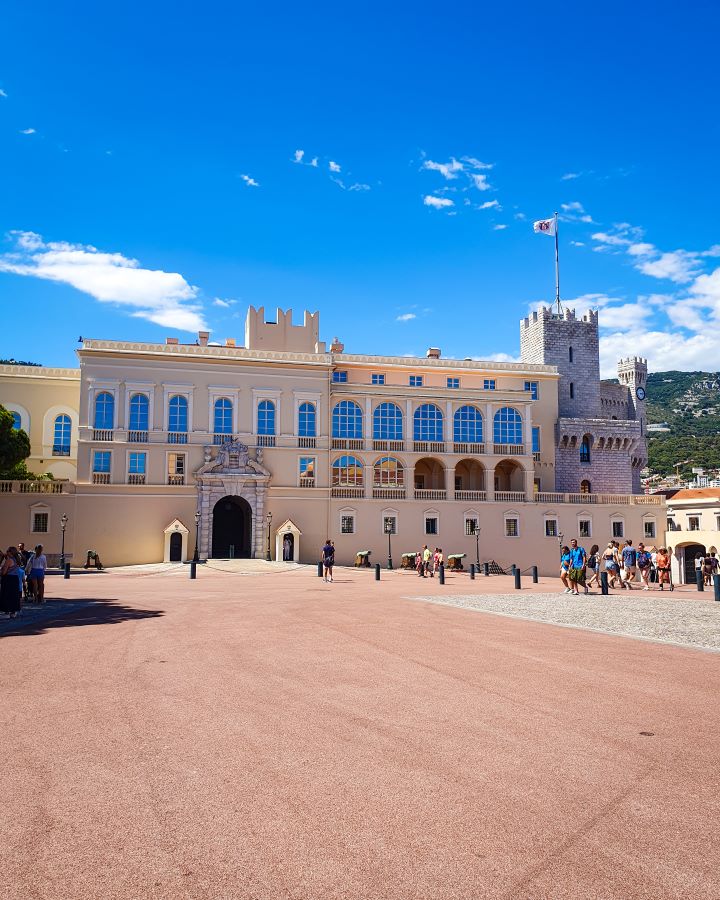 The Prince's Palace in Monaco. The building is sand coloured and not too big with a little turret on the right side with a flagpole on it. There's people out the front walking around the town square and the sky above is blue. "The Best Budget Nice to Monaco Day Trip"