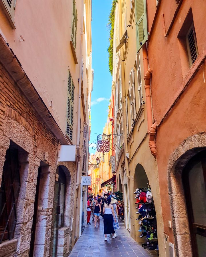 A narrow street in Monaco City. The walls on either side are tall and orange with windows and stone doorways. There's people walking around and little stalls too. "The Best Budget Nice to Monaco Day Trip"