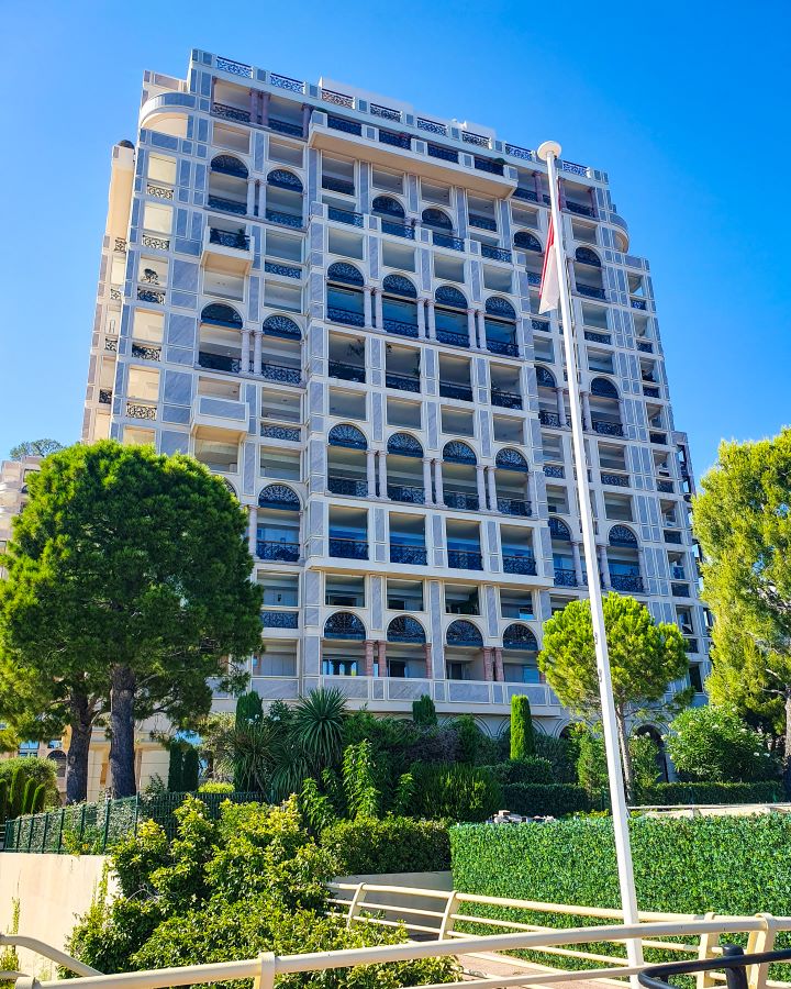 Another border from France to Monaco. There's a flag pole with the Monaco flag on it with a huge white hotel behind it with lots of black windows. There's green trees and bushes around and a blue sky above. "The Best Budget Nice to Monaco Day Trip"