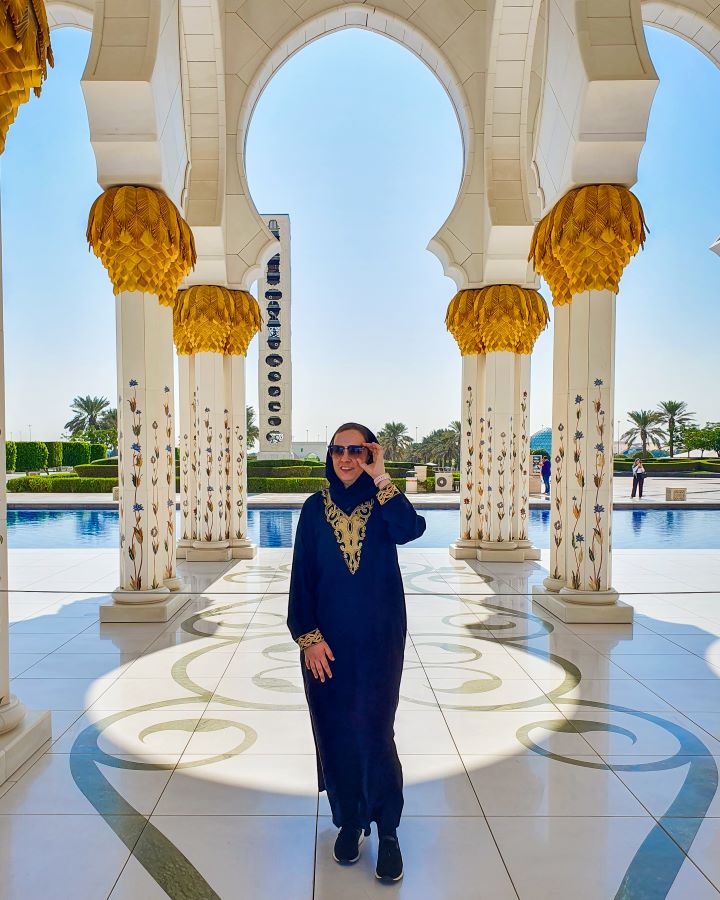 Abu Dhabi UAE Sheikh Zayed Grand Mosque from Dubai: Get Your Guide Review Krissie standing on a white marble floor with green patterns on it at the mosque in Abu Dhabi, UAE. She's wearing a black and gold abaya with sunglasses on. Behind her are 6 pillars with floral patterns and big gold bits on them and behind them is a blue pool and green plants. Sheikh Zayed Grand Mosque from Dubai: Get Your Guide Review