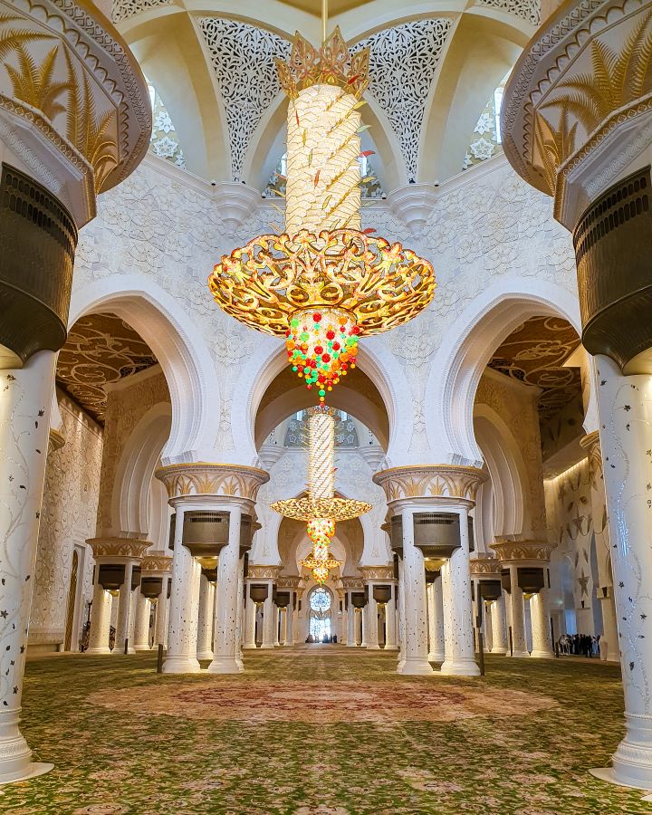 Abu Dhabi UAE Sheikh Zayed Grand Mosque from Dubai: Get Your Guide Review A big indoor hall in the mosque in Abu Dhabi, UAE with lots of pillars and a HUGE chandelier above with orange, red and green spots on it, however it's mostly made of gold. Under that is a carpet spreading the whole length of the room that's green, yellow and red. Sheikh Zayed Grand Mosque from Dubai: Get Your Guide Review