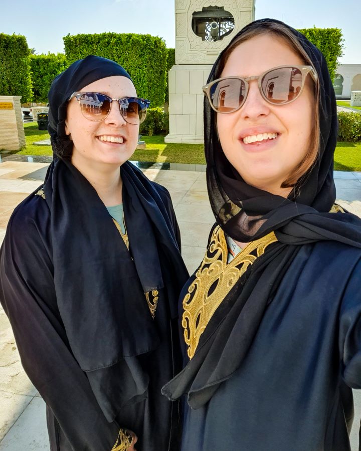 Abu Dhabi UAE Sheikh Zayed Grand Mosque from Dubai: Get Your Guide Review Krissie and Katherine smiling and looking at the camera at the mosque in Abu Dhabi, UAE. They're wearing sunglasses and have black and gold abayas on. Behind them are green plants and some bits of marble. Sheikh Zayed Grand Mosque from Dubai: Get Your Guide Review.