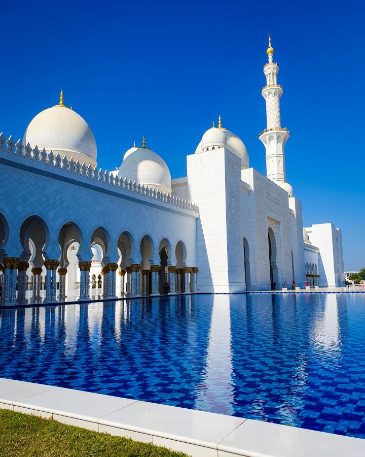 Abu Dhabi UAE Sheikh Zayed Grand Mosque from Dubai: Get Your Guide Review The outside of the white mosque in Abu Dhabi, UAE with a blue pool in front with a chequered pattern on the floor of the pool. The Mosque behind has domes and a minaret tower, all with gold spikes on top. Sheikh Zayed Grand Mosque from Dubai: Get Your Guide Review.