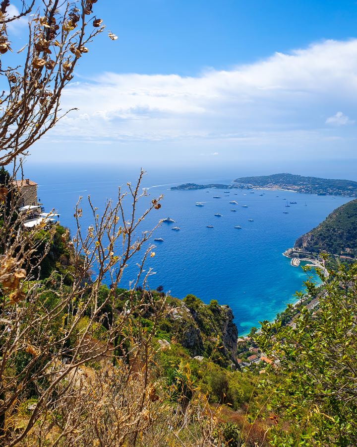 A view over the French Riviera in France. In the foreground are green and brown plants and there's a great view of the coastline with blue water. There's boats on the water and the sky above is cloudy and blue. How to Have the Best Three Days in Nice, France