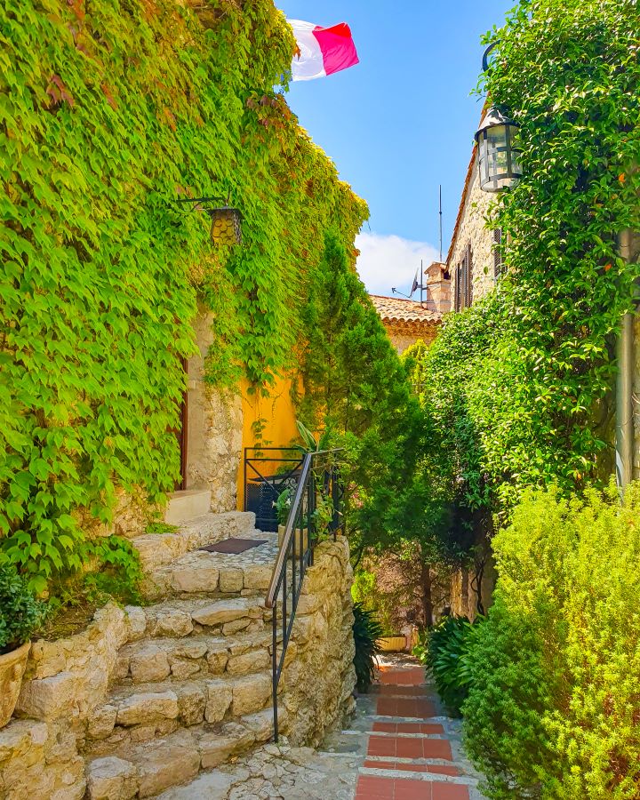 Inside Èze in France. On the left is a vine covered stone building with a French flag on it. There's a little path, more stone buildings with vines and a lamp on the right and the blue sky in the background. How to Have the Best Three Days in Nice, France