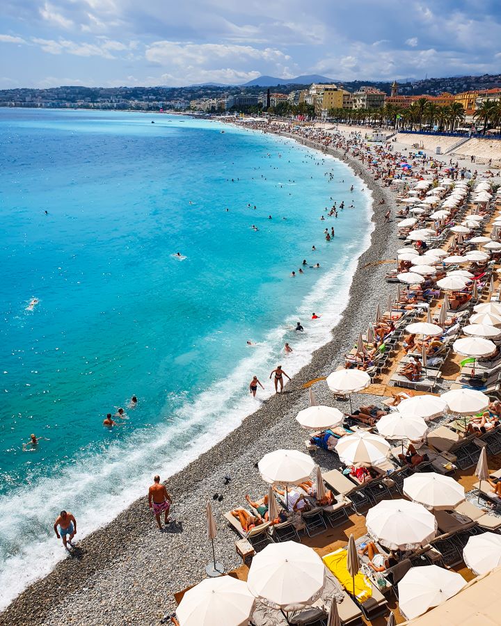 A long pebbly beach in Nice, France. On the right are all the sun lounges with white umbrellas over them and on the left is the blue water. In the background are coloured buildings, the hills and there's lots of people around. How to Have the Best Three Days in Nice, France