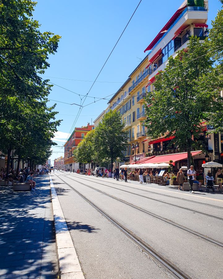 A long shopping street in Nice, France. On both sides of the street are big coloured buildings with trees out the front. Down the whole street are tram tracks with a wire overhead and there's people walking around as it's a nice sunny day. How to Have the Best Three Days in Nice, France