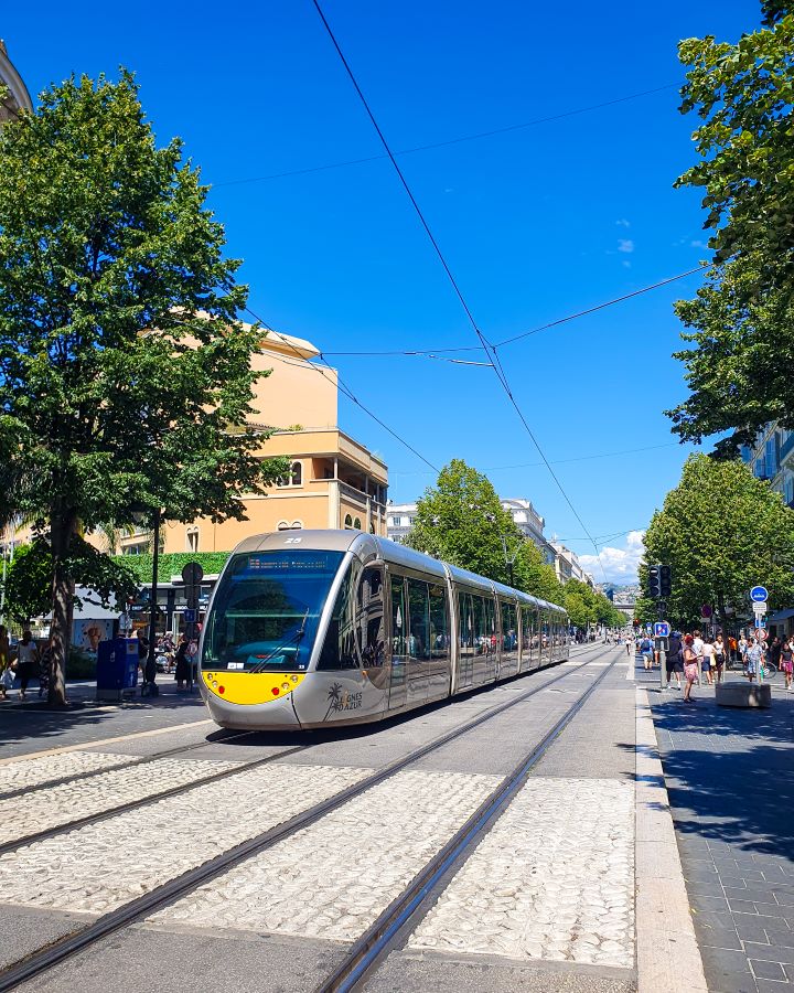 A silver tram with a yellow bit on the front going down some tracks on Avenue Jean Médecin in Nice, France. On either side of the tracks are streets with people walking down them, trees and shops. How to Navigate Public Transport in and Around Nice, France