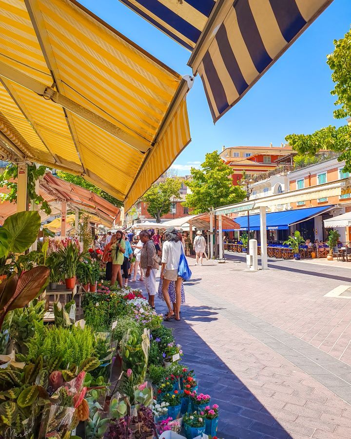 A little market in Nice, France. On the left are plants with coloured awnings overhead. There's people looking at the stalls and on the right are more coloured shops and a blue sky overhead. How to Have the Best Three Days in Nice, France
