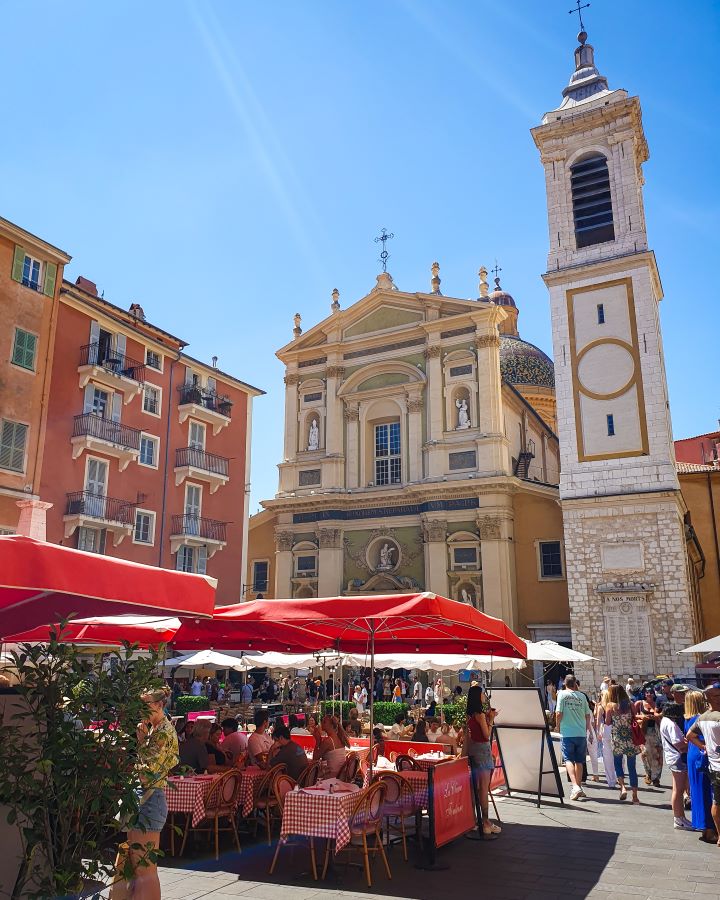 Inside Place Rossetti in Nice old town. There are red umbrellas with outdoor tables and plants with people sitting at them. Behind the tables is a big church with a tower next to it and some orange buildings next to it as well as people walk around the square. How to Navigate Public Transport in and Around Nice, France