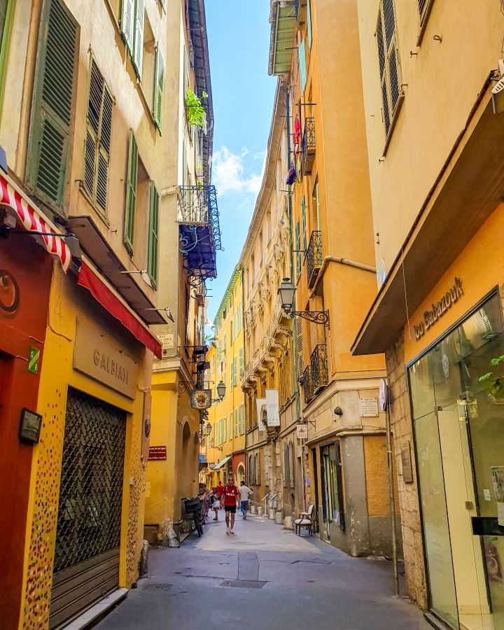 A little narrow street in Nice, France. The buildings on both sides are tall and a pale orange colour with cute window shutters, balconies and stores in the buildings. There's also some people walking down the street. How to Have the Best Three Days in Nice, France