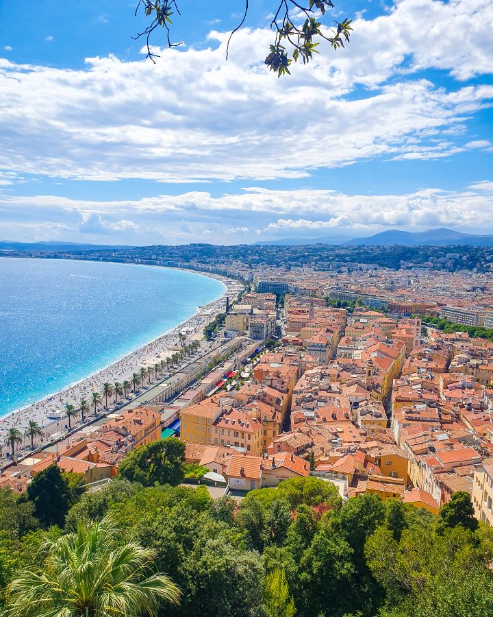 An amazing view over Nice, France from Castle Hill. In the foreground are some green trees with the orange of the city on the right and the blue water on the left. Behind those are some hills with the blue sky above it all. How to Have the Best Three Days in Nice, France