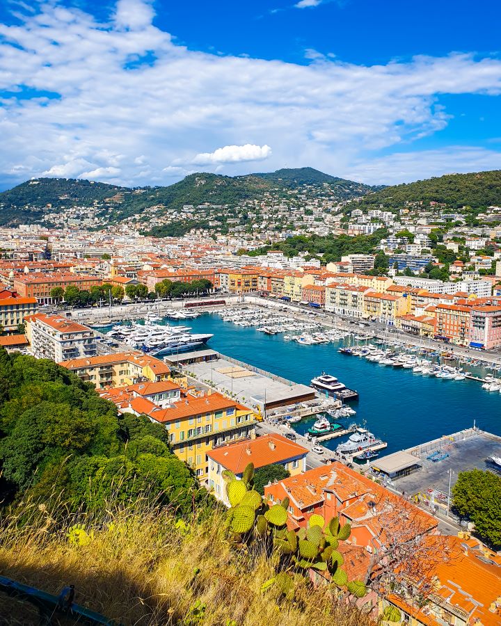 Looking over Quartier du port which is a marine harbour with lots of white boats docked there. There's also orange and yellow buildings in front of green hills. How to Navigate Public Transport in and Around Nice, France