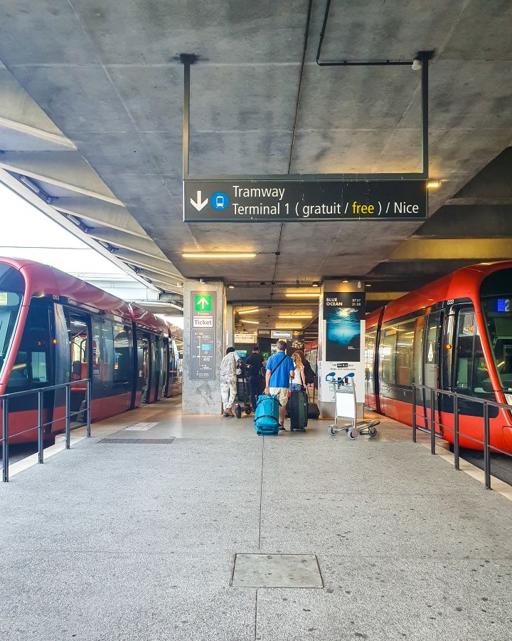 A tram terminal at the airport in Nice, France. There's two red and black trams with signs around in both French and English with some people with suitcases walking around too. How to Navigate Public Transport in and Around Nice, France