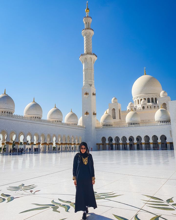 Abu Dhabi UAE Sheikh Zayed Grand Mosque from Dubai: Get Your Guide Review Katherine standing in the courtyard of the mosque in Abu Dhabi, UAE with coloured leaf patterns on the ground. Behind her is a walkway with arches in it, and lots of white domes and one minaret, all with gold spikes on top. Sheikh Zayed Grand Mosque from Dubai: Get Your Guide Review.