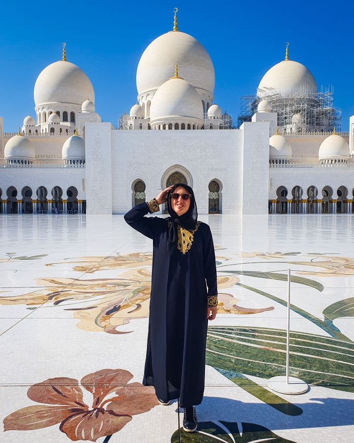 Abu Dhabi UAE Sheikh Zayed Grand Mosque from Dubai: Get Your Guide Review Sheikh Zayed Grand Mosque from Dubai: Get Your Guide Review. Katherine standing in a marble courtyard in the mosque in Abu Dhabi, UAE with coloured flowers on the ground and the rest is white. Behind her are 3 big white domes, with a fourth smaller dome too. All 4 domes have gold spikes on them and the sky behind is clear and blue.