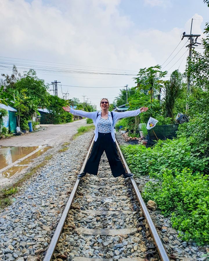 Maeklong Railway market Thailand Floating and Train Markets from Bangkok: Get Your Guide Review Katherine standing and posing on some train tracks in Thailand. Under the tracks are rocks, on the the right are green plants and on the left is a road with more plants next to it. Floating and Train Markets from Bangkok: Get Your Guide Review