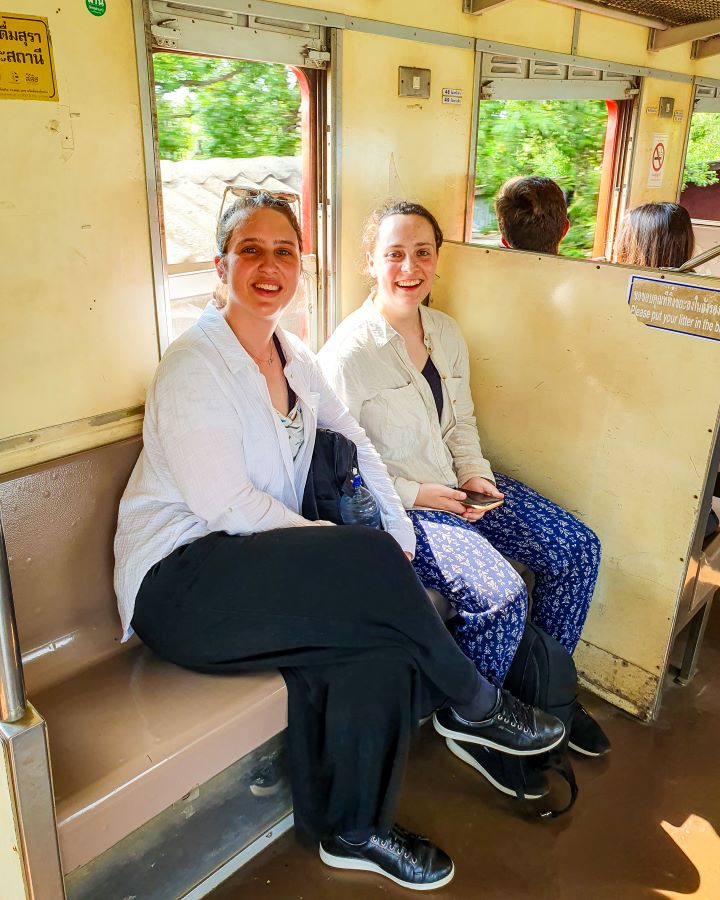 Maeklong Railway market Thailand Floating and Train Markets from Bangkok: Get Your Guide Review Krissie and Katherine sitting on a seat on a train in Thailand. They're wearing long pants and have their hair up and you can see trees passing by the train behind them. Floating and Train Markets from Bangkok: Get Your Guide Review