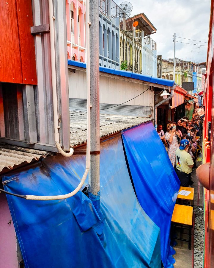 Maeklong Railway market Thailand Floating and Train Markets from Bangkok: Get Your Guide Review Going through the Maeklong Railway market in Thailand on a train. There's blue awnings that have been packed away as the train goes through. There's lots of buildings next to the train with people standing out the front taking photos of the train. Floating and Train Markets from Bangkok: Get Your Guide Review
