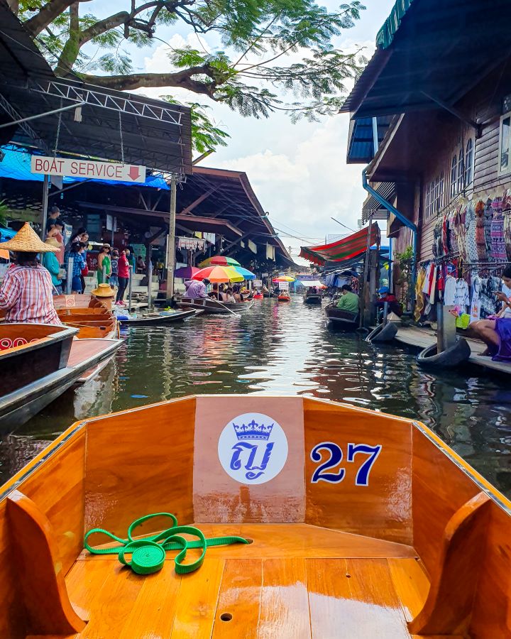 Damnoen Saduak floating market Thailand Floating and Train Markets from Bangkok: Get Your Guide Review Floating and Train Markets from Bangkok: Get Your Guide Review. The front of a little wooden boat with a blue 27 on it at a floating market in Thailand. In front of the boat is water with land on either side with market stalls selling colourful items with people all around selling and buying stuff.