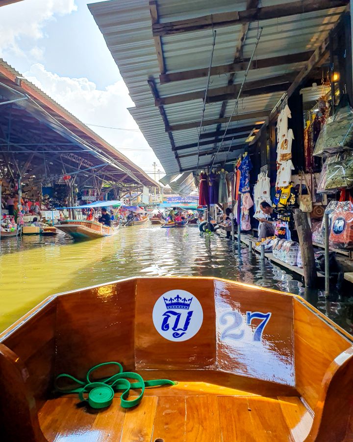 Damnoen Saduak floating market Thailand Floating and Train Markets from Bangkok: Get Your Guide Review On a little wooden boat with a blue 27 on the inside at the Damnoen Saduak floating market in Thailand. Around the boat are stalls on the land with awnings over them and people selling clothes and fresh food. Floating and Train Markets from Bangkok: Get Your Guide Review