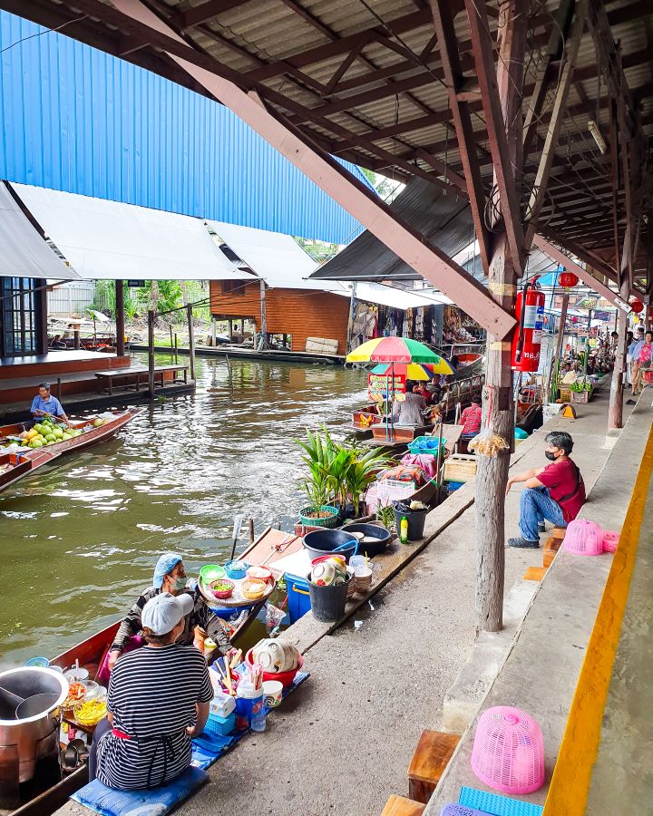 Damnoen Saduak floating market Thailand Floating and Train Markets from Bangkok: Get Your Guide Review Sitting on the land looking out at the Damnoen Saduak floating market in Thailand. There's people sitting on some concrete stairs and in the boats selling fresh food and souvenirs. There's water with more wooden boats on it with awnings over it and more stalls in the background. Floating and Train Markets from Bangkok: Get Your Guide Review