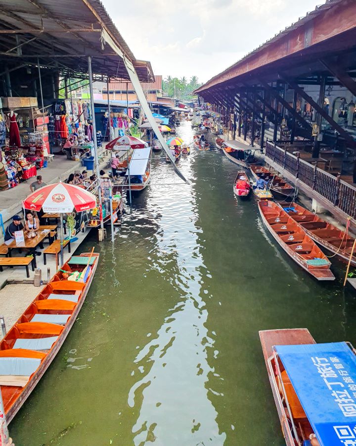 Damnoen Saduak floating market Thailand Floating and Train Markets from Bangkok: Get Your Guide Review A floating market in Thailand. There's brown water going down the middle with little wooden boats on either side. Next to them is land with little stalls, tables and chairs, and people on them. Floating and Train Markets from Bangkok: Get Your Guide Review