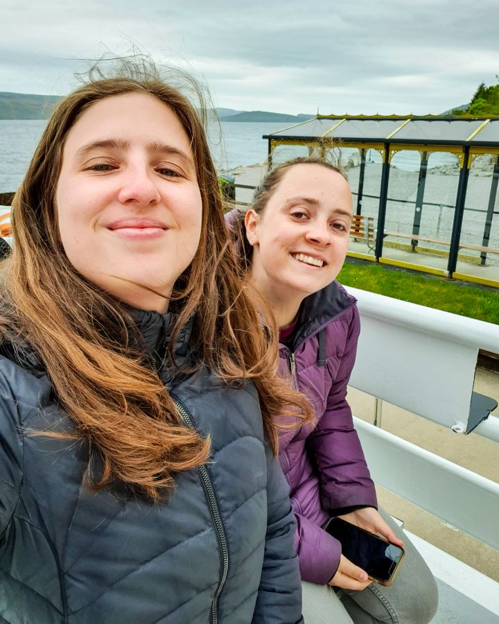 Urquhart Castle Loch Ness Scotland Loch Ness Cruise From Inverness: Get Your Guide Review Loch Ness Cruise From Inverness: Get Your Guide Review. Katherine and Krissie wearing puffer jackets and smiling at the camera. They're on a boat and behind them is a little undercover seated area on the land, and you can see a bit of Loch Ness behind too in Scotland.
