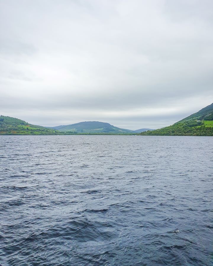 Urquhart Castle Loch Ness Scotland Loch Ness Cruise From Inverness: Get Your Guide Review On the water as we cross Loch Ness in Scotland. The water is a grey blue and behind it are green hills and an overcast sky. Loch Ness Cruise From Inverness: Get Your Guide Review