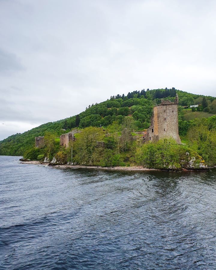 Urquhart Castle Loch Ness Scotland Loch Ness Cruise From Inverness: Get Your Guide Review The blue water of the Loch with the stone ruins of Urquhart Castle in the background in Scotland. There's green trees all around the castle and a green hill behind it. Loch Ness Cruise From Inverness: Get Your Guide Review