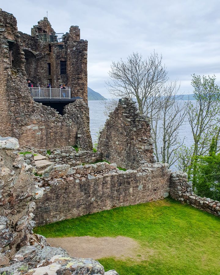 Urquhart Castle Loch Ness Scotland Loch Ness Cruise From Inverness: Get Your Guide Review Stone ruins of Urquhart Castle in Scotland. There's a platform up on the second level of a ruin with a white railing and people looking out from it. There's also a patch of green grass and you can see some of the Loch and hills in the background. Loch Ness Cruise From Inverness: Get Your Guide Review