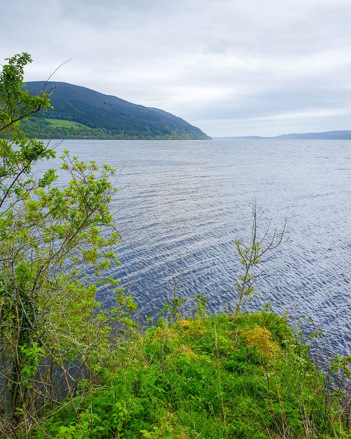 Urquhart Castle Loch Ness Scotland Loch Ness Cruise From Inverness: Get Your Guide Review Some green trees in the foreground with the moody blue Loch in the background in Scotland. Behind that are some dark green hills and the overcast sky. Loch Ness Cruise From Inverness: Get Your Guide Review
