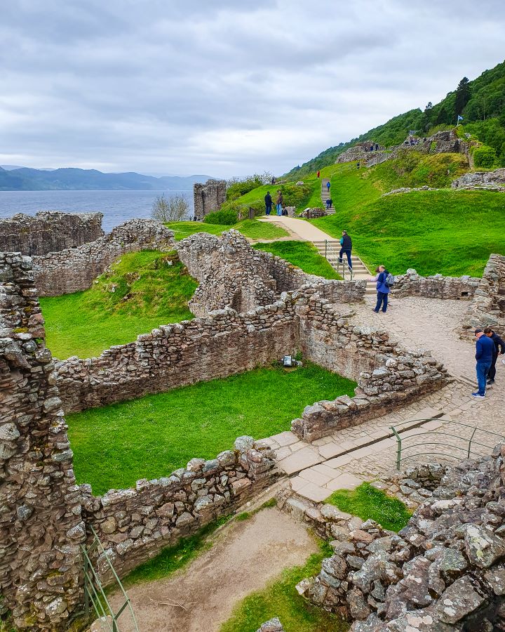 Urquhart Castle Loch Ness Scotland Loch Ness Cruise From Inverness: Get Your Guide Review Some dirt paths running through the stone ruins of Urquhart Castle in Scotland. There's people walking around, patches of green grass and green trees and you can see the moody blue Loch and hills behind it all. Loch Ness Cruise From Inverness: Get Your Guide Review