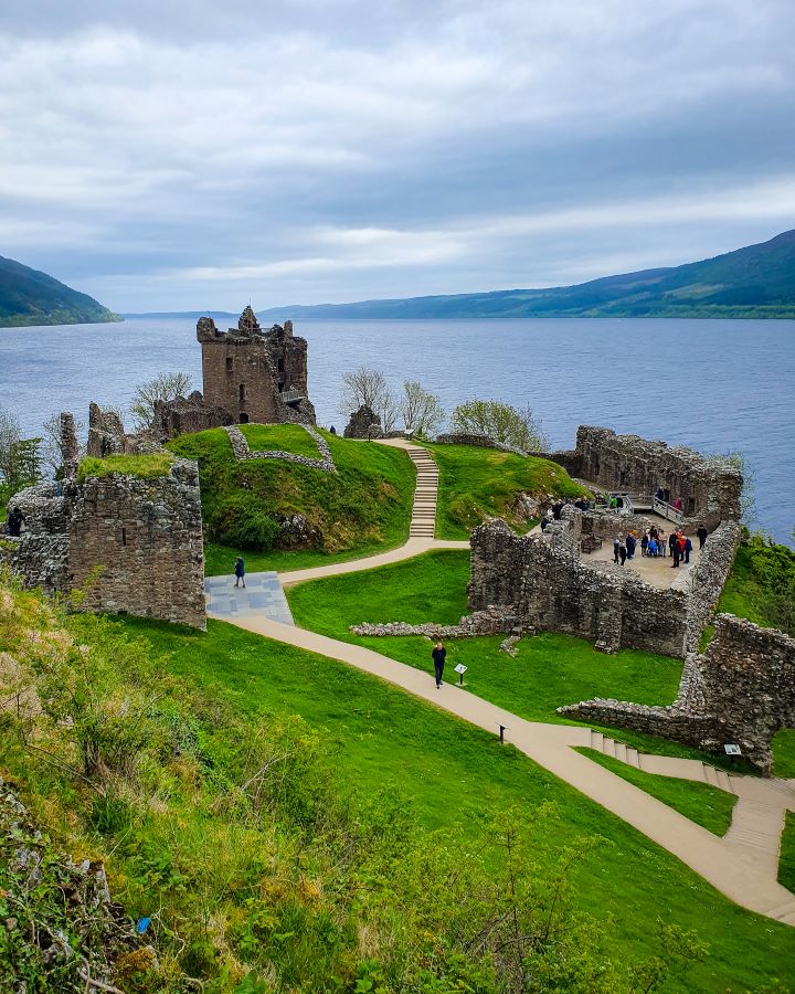 Urquhart Castle Loch Ness Scotland Loch Ness Cruise From Inverness: Get Your Guide Review Looking over Urquhart Castle in Scotland. In the foreground is green grass and behind that are paths going through stone ruins of the castle. There's people walking around the ruins and behind the castle is the blue Loch. You can also see some dark green hills on either side of the Loch. Loch Ness Cruise From Inverness: Get Your Guide Review