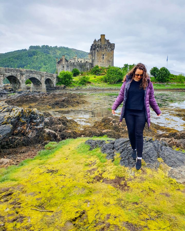 Krissie standing on a rock with her purple puffer jacket on in front of Eilean Donan Castle in Scotland. Behind her is a little stone castle with a stone bridge going over to the island it's on and there's also green trees and grass on the island and on another hill behind it. Isle of Skye From Inverness: Get Your Guide Review