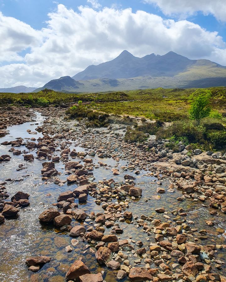 A beautiful stop along the way to the Isle of Skye, Scotland. In the foreground is a river with rocks and behind it is green grass and a huge pointy hill under a cloudy sky. Isle of Skye From Inverness: Get Your Guide Review