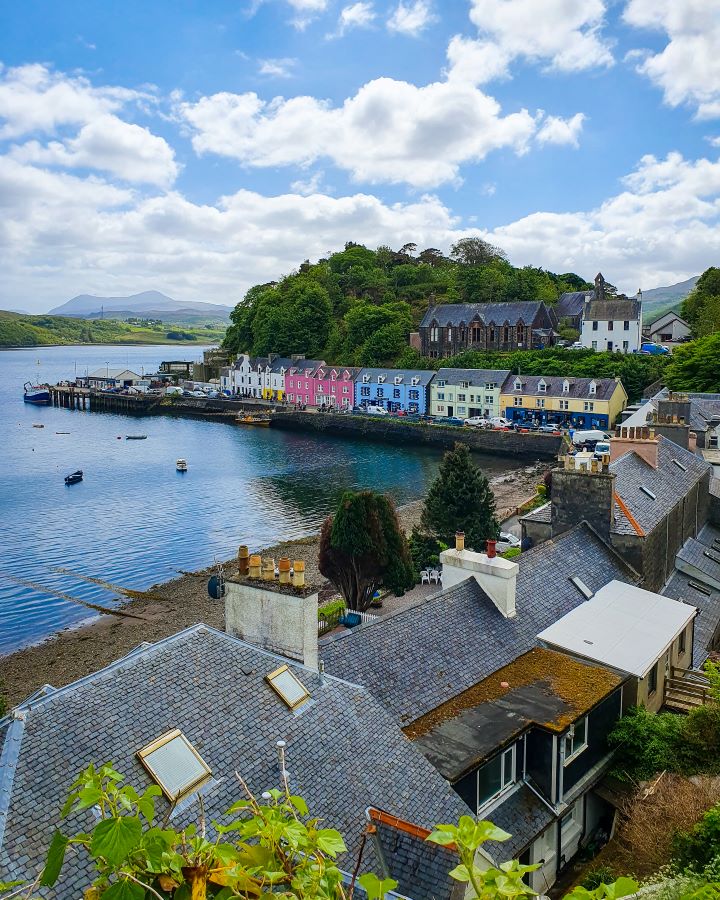 Looking over Portree in the Isle of Skye, Scotland. In the foreground there's little houses with grey roofs, behind them is a little beach with coloured buildings alongside it with pink, blue and yellow houses. Behind the coloured buildings is a green hill and in the background are more hills. Isle of Skye From Inverness: Get Your Guide Review