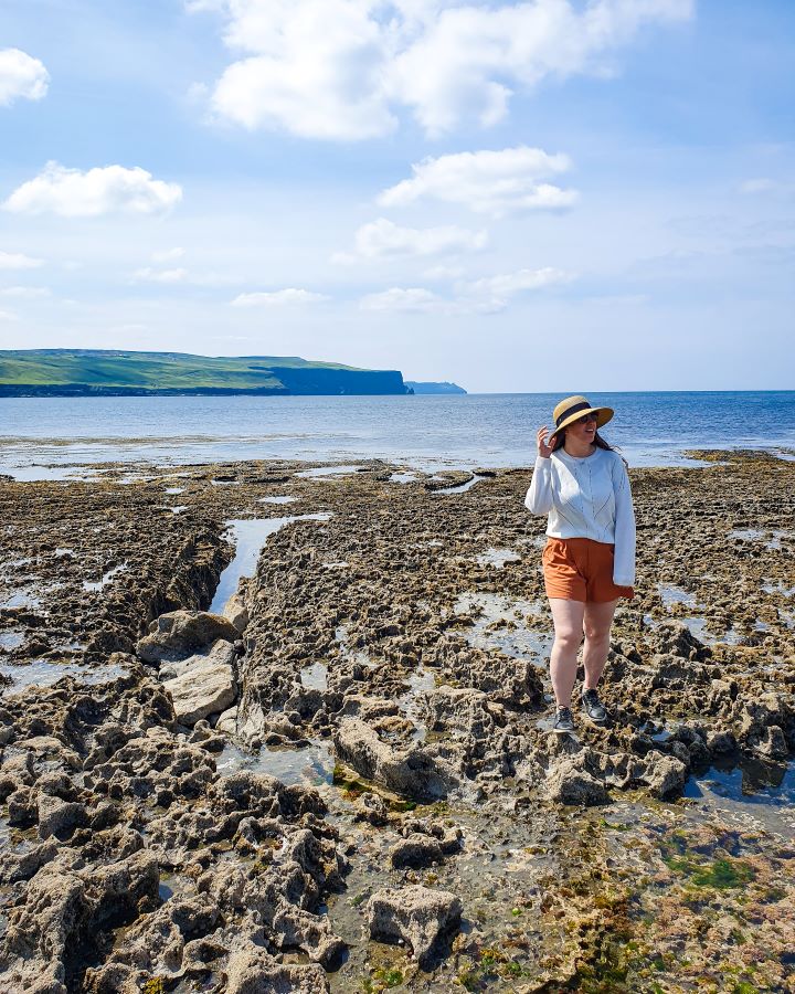 Ireland Cliffs of Moher From Galway: Get Your Guide Review Katherine standing on some rocks beside the Atlantic ocean in Ireland. Behind her is the ocean and some green cliffs. Cliffs of Moher From Galway: Get Your Guide Review