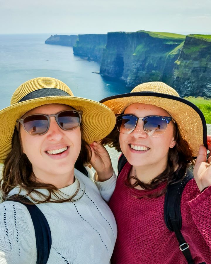Ireland Cliffs of Moher From Galway: Get Your Guide Review Krissie and Katherine smiling and holding their hats on their heads. Behind them are the amazing cliffs of Moher in Ireland. The cliffs are big with green grass on top and you can see the cliffs sprawling out along the water. Cliffs of Moher From Galway: Get Your Guide Review