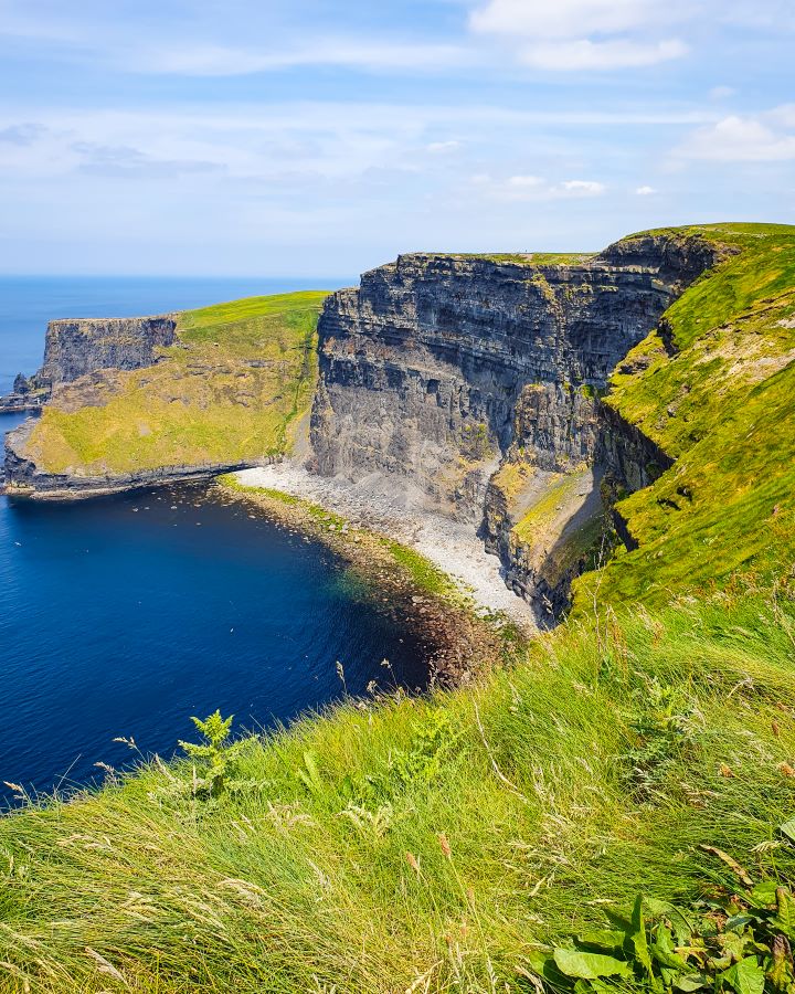Ireland Cliffs of Moher From Galway: Get Your Guide Review In the foreground is green grass and behind that is a big white cliff with green grass on top in Ireland. In front of the cliff is blue water with blue cloudy sky above it. Cliffs of Moher From Galway: Get Your Guide Review