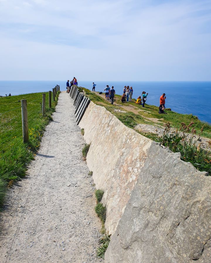 Ireland Cliffs of Moher From Galway: Get Your Guide Review A stone barrier with a pathway and grass on the left of it in Ireland. To the right of the barrier are lots of stupid people taking photos beside the cliff and you can see blue water in the background. Cliffs of Moher From Galway: Get Your Guide Review