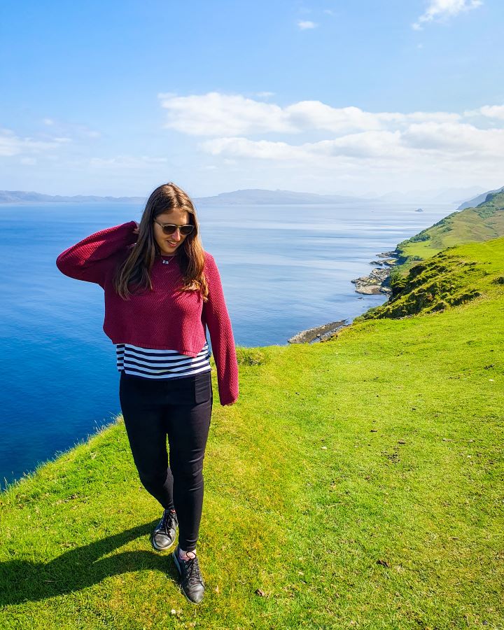 Isle of Skye From Inverness: Get Your Guide Review. Katherine standing on a green grassy cliff with clear blue water behind her. In the background you can see hills coming out of the water and the sky is blue and cloudy.