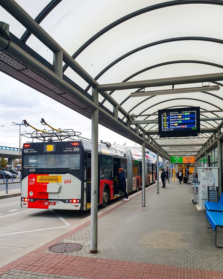 Prague Czechia A Complete Guide to Prague Public Transport A bus station at Prague airport, Czechia. On the right is the bus stop with places to sit and wait with a number 59 bus on the left. Above there's a roof in case it rains and the sky is overcast. A Complete Guide to Prague Public Transport